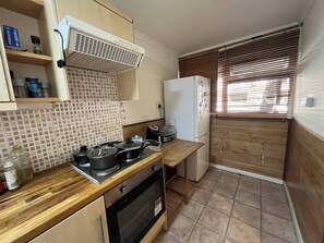 Eine kleine Küche mit Herd, Backofen und Kühlschrank. Ein Fenster mit Jalousien und eine geflieste Spritzschutzleiste sind vorhanden.