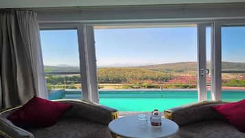 A room with a view of a pool and landscape, two armchairs, a small table with a bottle and glasses.