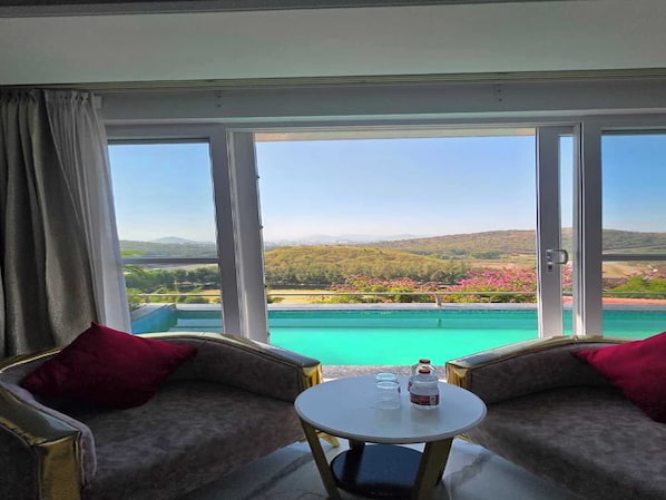 A room with a view of a pool and landscape, two armchairs, a small table with a bottle and glasses.