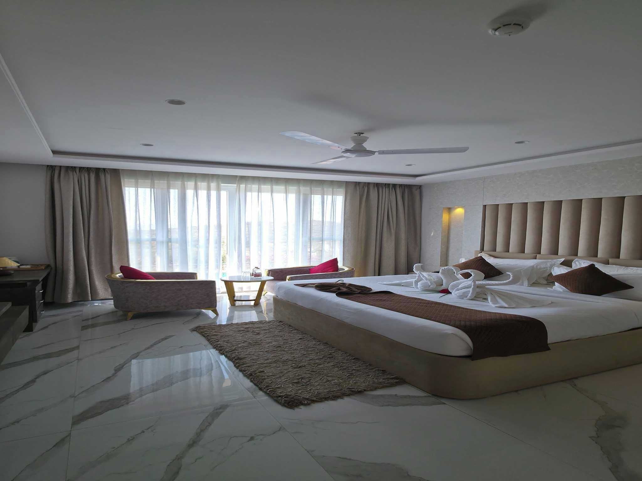 A hotel room with a large bed, a desk, a chair, a ceiling fan, and a view of the outside.