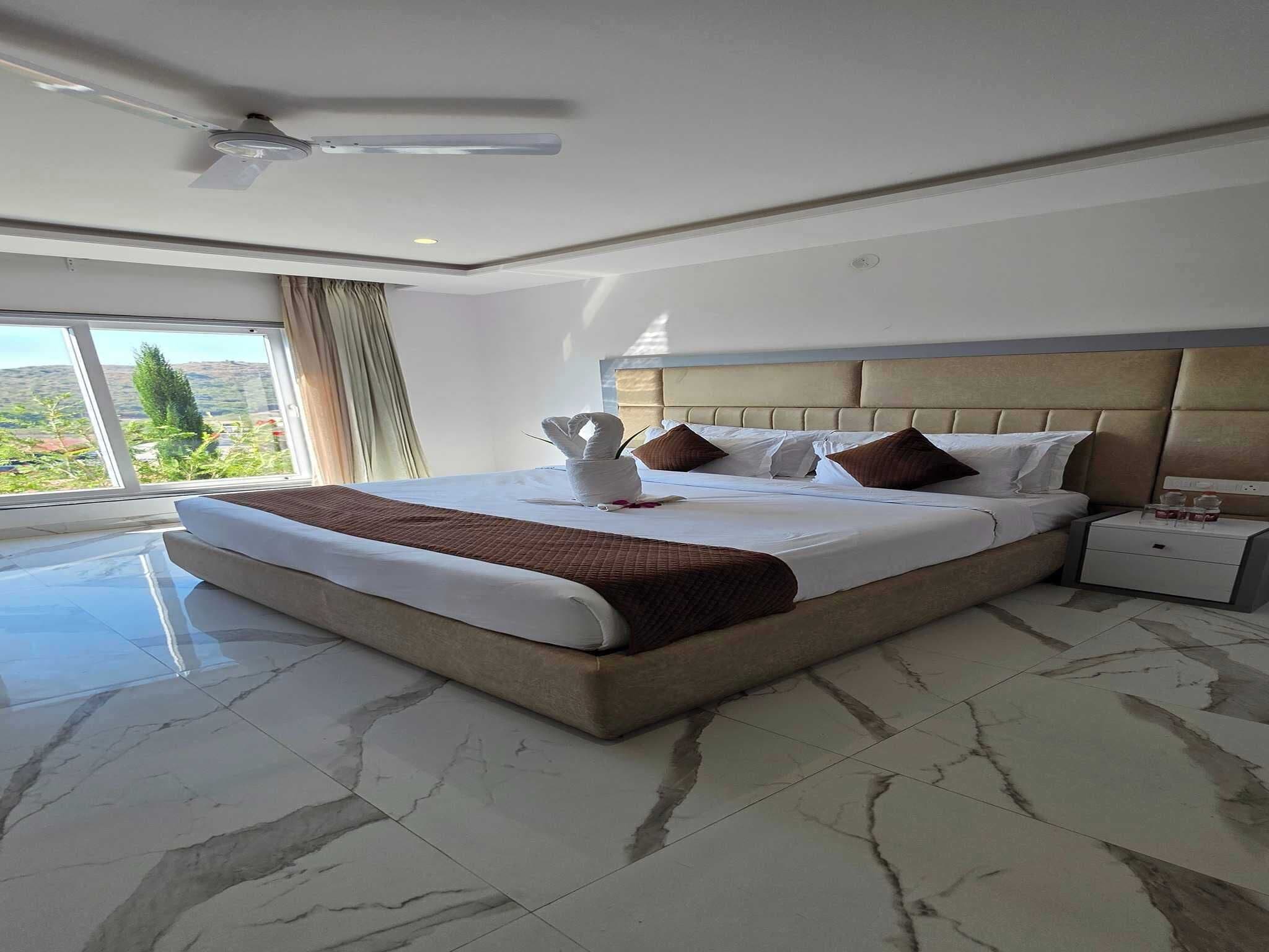 A hotel room with a large bed, a nightstand, a ceiling fan, and a view of the outdoors.