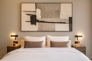 Ein Hotelzimmer mit einem großen, abstrakten Gemälde, zwei Nachttischen mit Lampen sowie einem Bett mit gestreifter Bettwäsche.