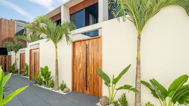 Um edifício moderno com entrada protegida por grades, cercado por plantas tropicais e um calçadão pavimentado.