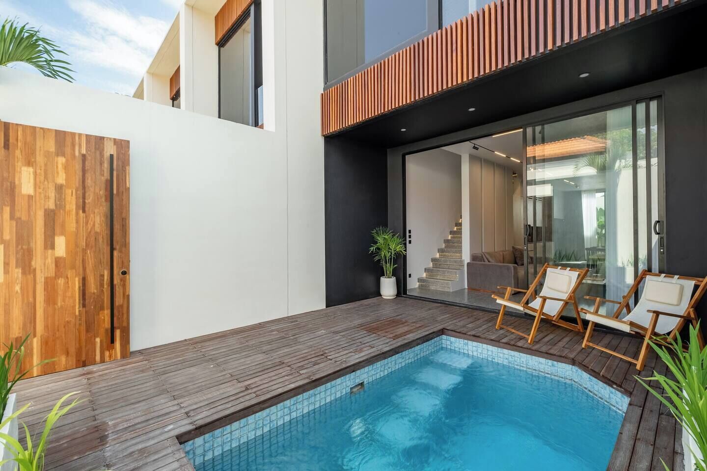 Un área de piscina moderna al aire libre con cubierta de madera, sillas de descanso y una puerta de cristal que da a una escalera.