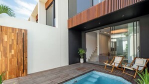 Uma área de piscina moderna ao ar livre, com deck de madeira, espreguiçadeiras e uma porta de vidro que dá para uma escadaria.