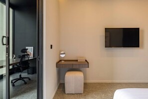 Ein Hotelzimmer mit Schreibtisch, Stuhl, Nachttisch, Spiegel und einem an der Wand befestigten Fernseher.
