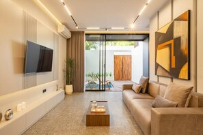 Ein modernes Wohnzimmer mit Fernseher, Couchtisch aus Holz, beiger Couch und Glastür zum Außenbereich mit Pool.