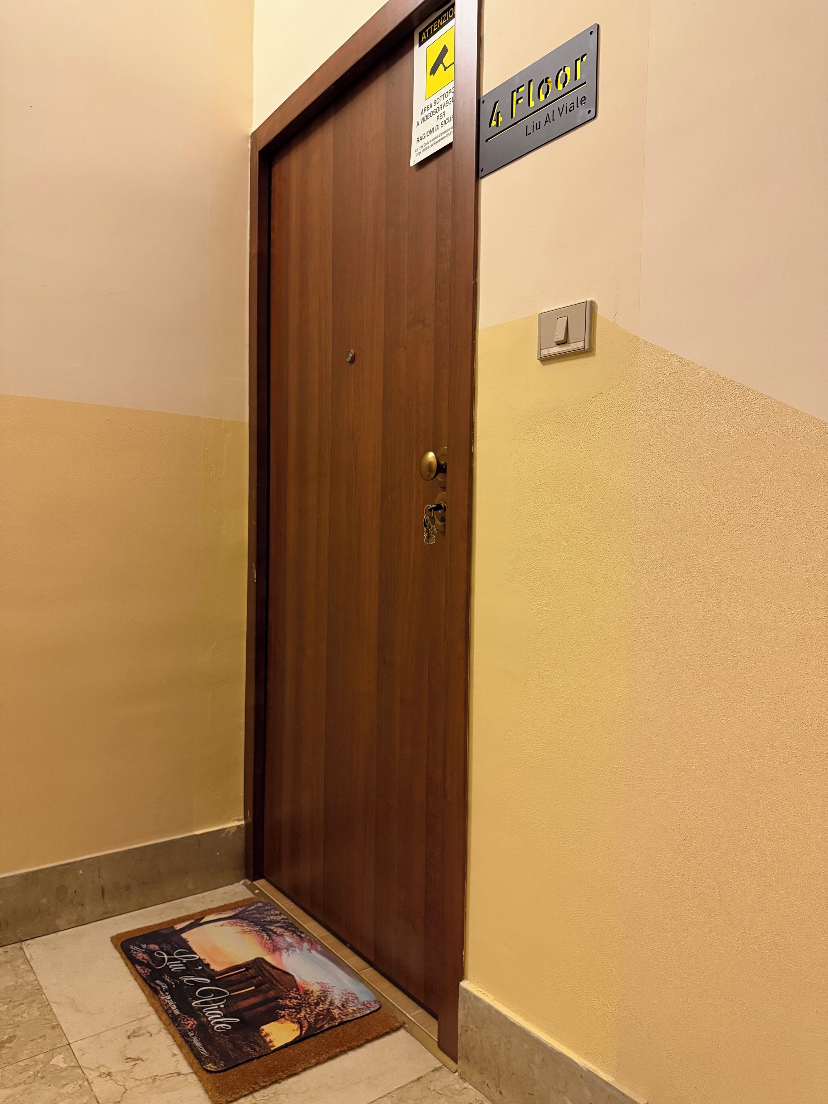 Porta de madeira com tapete de entrada exibindo uma imagem panorâmica. Uma placa acima da porta indica o número do quarto '4 ANDAR' e o nome 'Liu Ai Viale'.