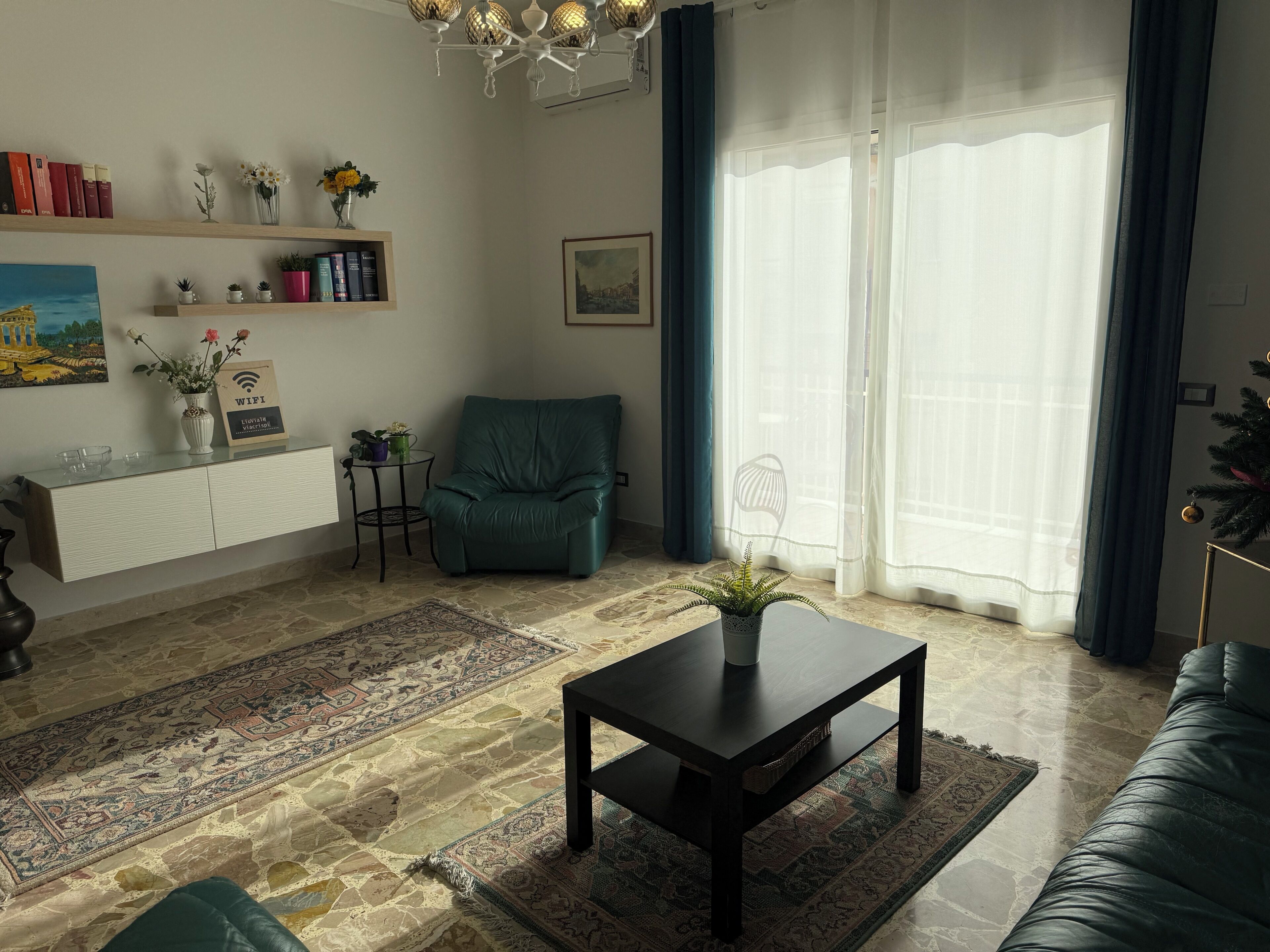 A living room with a sofa, a teal armchair, a coffee table, a bookshelf, and a large window with sheer curtains.