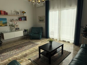 Uma sala de estar com sofá, uma poltrona verde-água, mesa de centro, estante de livros e uma janela grande com cortinas leves.