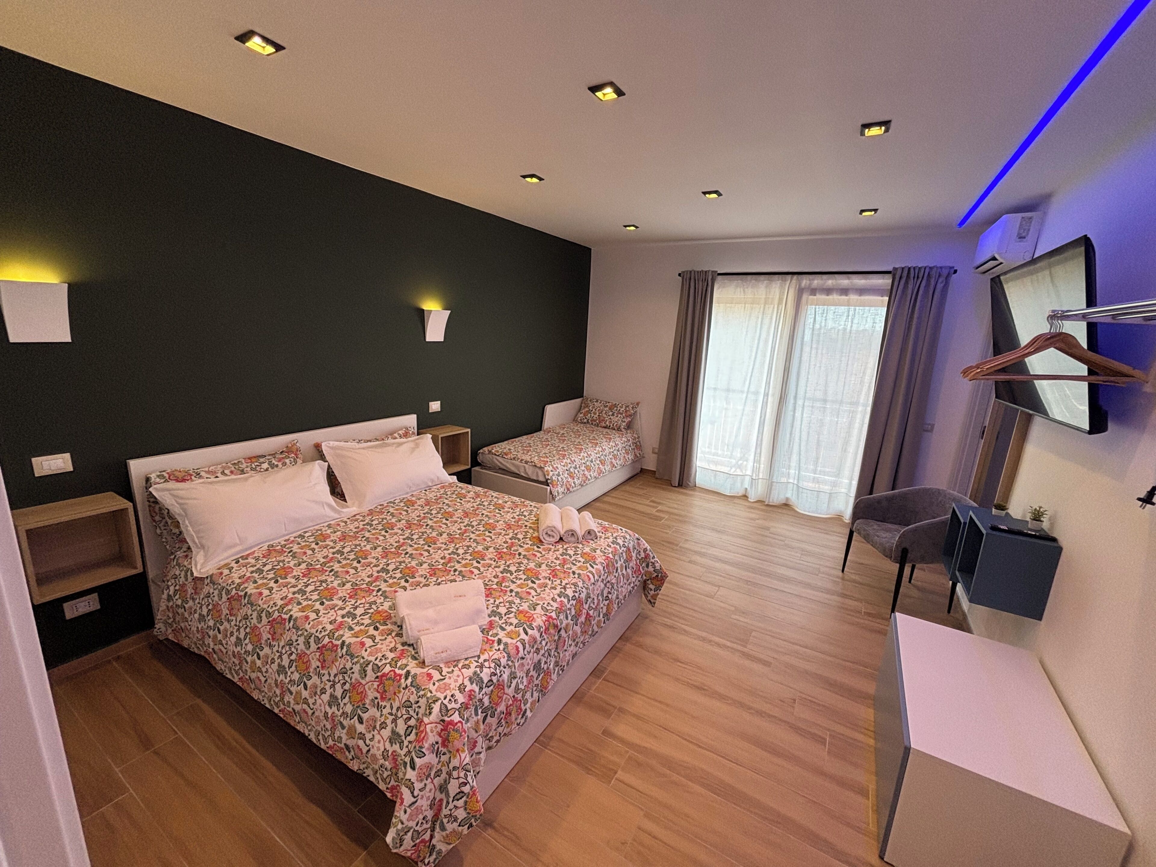 Une chambre d’hôtel avec un grand lit, un lit plus petit, un bureau, une chaise, une armoire et une fenêtre avec des rideaux.