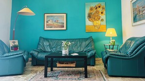 Un salón comedor color turquesa con un sofá, dos sillones, una mesa de centro con un jarrón de flores, una lámpara y cuadros en las paredes.