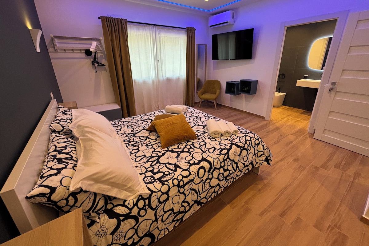 Un dormitorio moderno con cama, una silla, televisión y un baño visible a través de una puerta abierta.