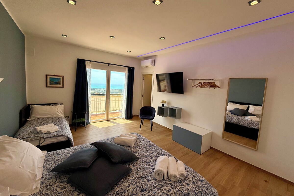 Habitación de hotel con balcón, cama con almohadas, escritorio con silla, televisor y espejo.