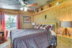 Een slaapkamer met een bed, een plafondventilator, een raam met gordijnen, een ingelijst schilderij en een bedlamp.