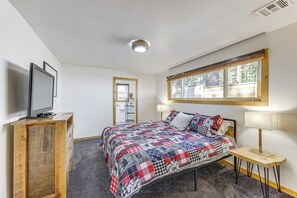 Quarto com cama, penteadeira de madeira, televisão, janela com persianas e porta para outro cômodo.