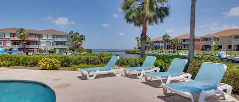 A pool area with lounge chairs, a clear view of the sea, and a row of multi-story buildings with balconies.