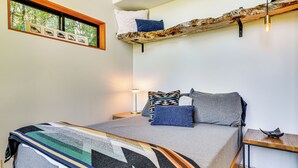 Una cama con una colcha estampada, almohadas, una mesita de noche con una lámpara y una ventana con vistas a árboles.