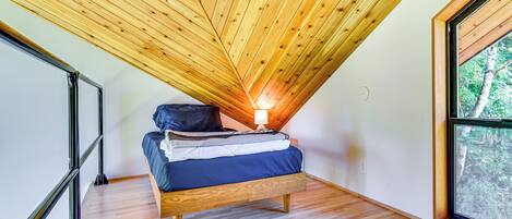 A bedroom with a sloped ceiling, a bed with pillows, a nightstand, and a window with a view of trees.