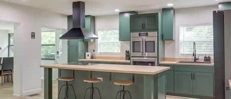 Una cocina moderna con una isla central, taburetes de bar, horno empotrado y un ventanal grande.