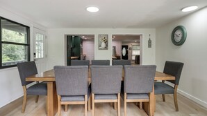 Een eethoek met een houten tafel en grijze fauteuils, een raam met uitzicht op groen, en een wandklok.