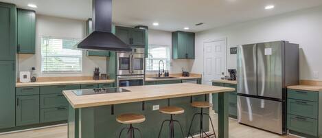 Una cocina moderna con armarios verdes, un refrigerador de acero inoxidable, una isla central con taburetes y una campana extractora negra.