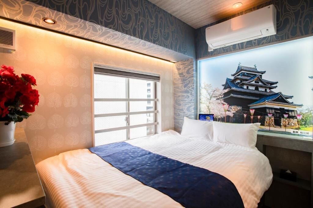 A hotel room with a large bed, a window with blinds, a wall-mounted air conditioner, and a decorative wall with a traditional building illustration.