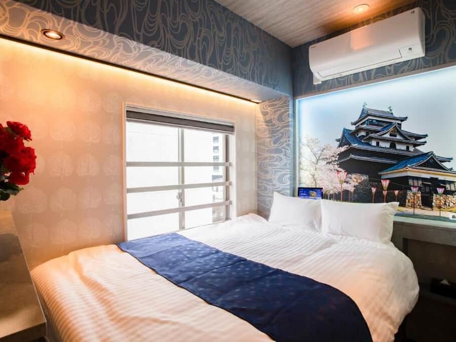 Habitación de hotel con una cama grande, una ventana con persianas, un aire acondicionado instalado en la pared y una decoración mural con una ilustración de una construcción tradicional.