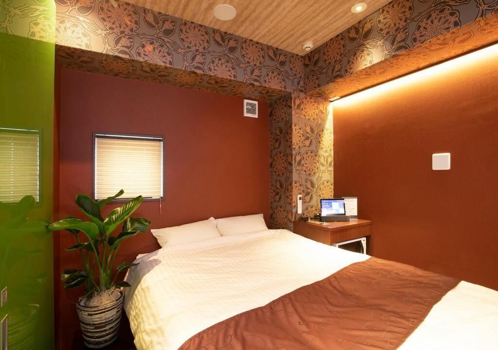 Een hotelkamer met een bed, een nachtkastje met een lamp, een kamerplant en een raam met jaloezieën.