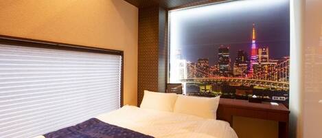 Habitación de hotel con cama, ventana con vista a la ciudad, escritorio y lámpara de pared.