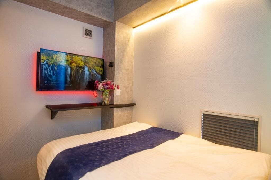 Une chambre d’hôtel avec un lit, un téléviseur fixé au mur, une étagère avec des fleurs et un climatiseur fixé au mur.