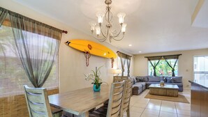 Een eetgedeelte met een houten tafel en stoelen, een surfboard als decoratie, een kroonluchter, en een zithoek met een bank en salontafel.