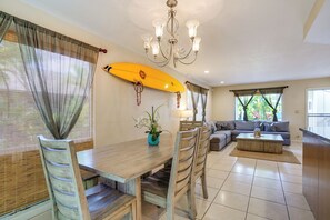 A dining area with a wooden table and chairs, a yellow surfboard decoration, a chandelier, and a living area with a sofa and coffee table. - Walk to Sunset Beach! Extended Escape w/ Patio (Haleiwa)