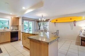 A kitchen with a central island, wooden cabinets, a dishwasher, and a surfboard mounted on the wall. - Walk to Sunset Beach! Extended Escape w/ Patio (Haleiwa)