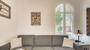En stue med en grå hjørnesofa, puder med mønster, et vintageskilt, et vindue med persienner og et lille træ sidebord med en lampe.