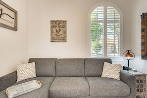 Ein Wohnzimmer mit einem grauen Ecksofa, gemusterten Kissen, einem Vintage-Schild, einem Fenster mit Jalousien und einem kleinen Holztisch mit Lampe.