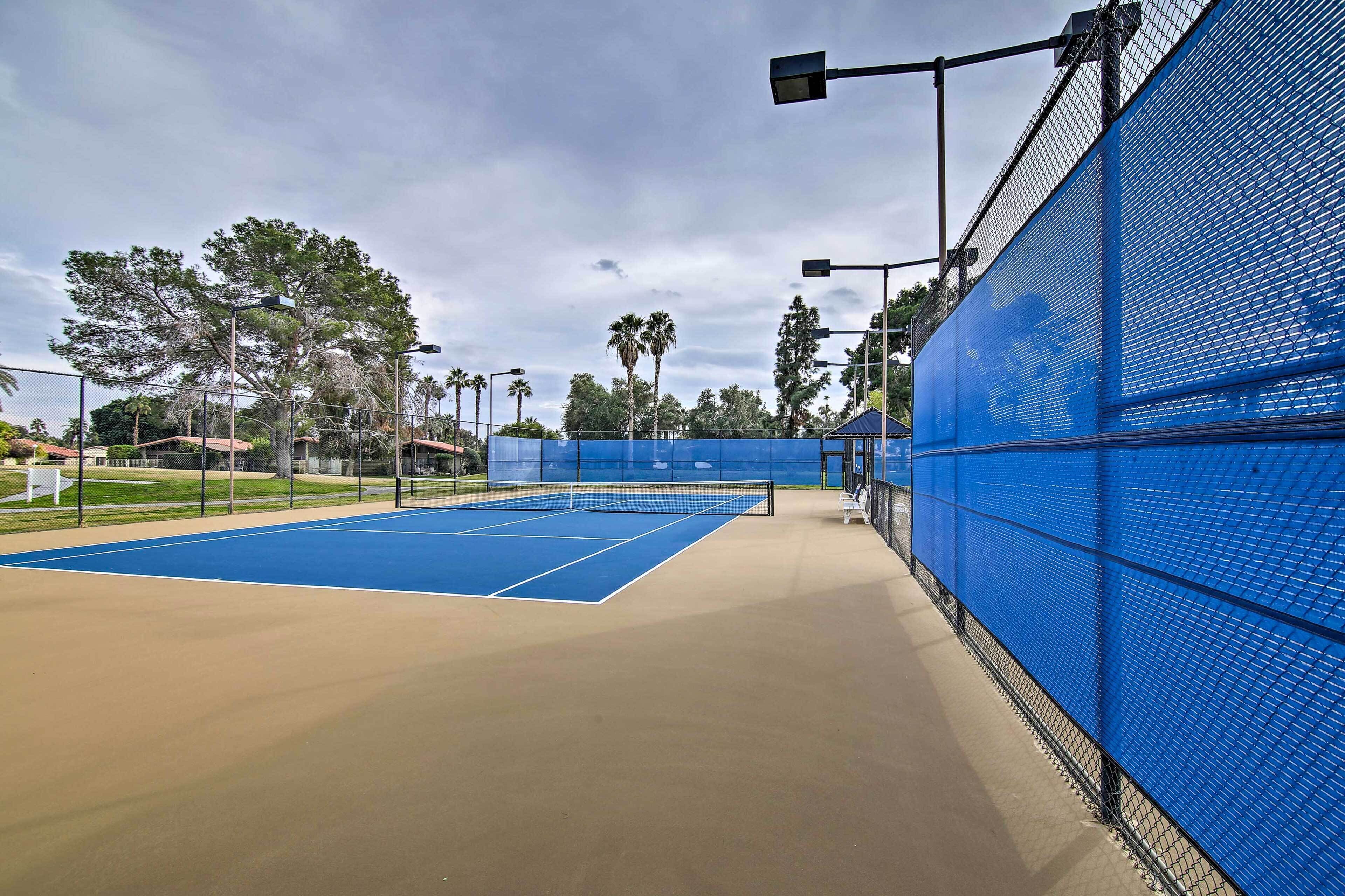 Ein Tennisplatz mit blauer Oberfläche und einem beigefarbenen Umfeld, umgeben von einem blauen Zaun und Straßenlaternen.