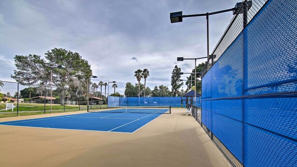 En tennisbane med blå overflade og et beige område omkring, omgivet af et blåt hegn og gadelamper.