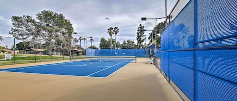Een tennisbaan met een blauwe oppervlakte en een beige omliggend gebied, omringd door een blauw hek en lantaarnpalen.