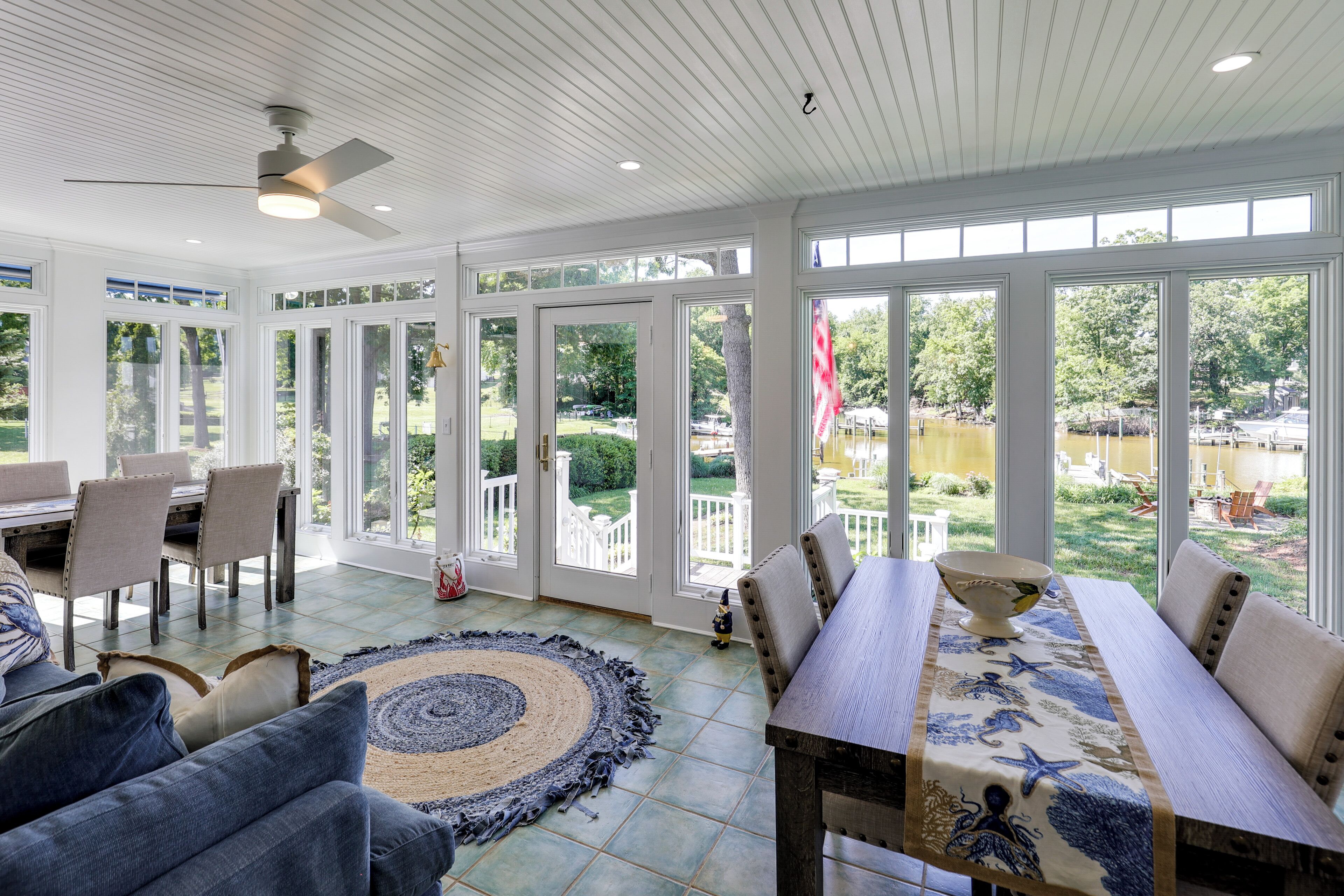 Una habitación amplia con comedor, ventilador de techo y grandes ventanas que ofrecen vistas a un jardín y mobiliario exterior.