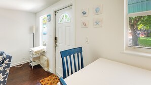 Ein Zimmer mit weißer Tür, einem Fenster mit Blick auf Grünflächen, einem weißen Tisch, einem blauen Stuhl und einem gemusterten Teppich.