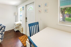 A room with a white door, a window with a view of greenery, a white table, a blue chair, and a patterned rug. - Walkable Dtwn Spot! Bright Bungalow Near Beach (Lake Worth Beach)