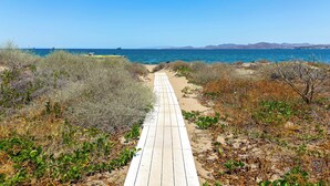 Una pasarela de madera que lleva a una playa con aguas azules cristalinas y montañas a lo lejos.