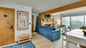 A cozy living room with a blue sofa, wooden flooring, a fireplace, and a large window overlooking a balcony. - Near Slopes, Golf, Lake! Shanty Creek Resort Condo (Bellaire)