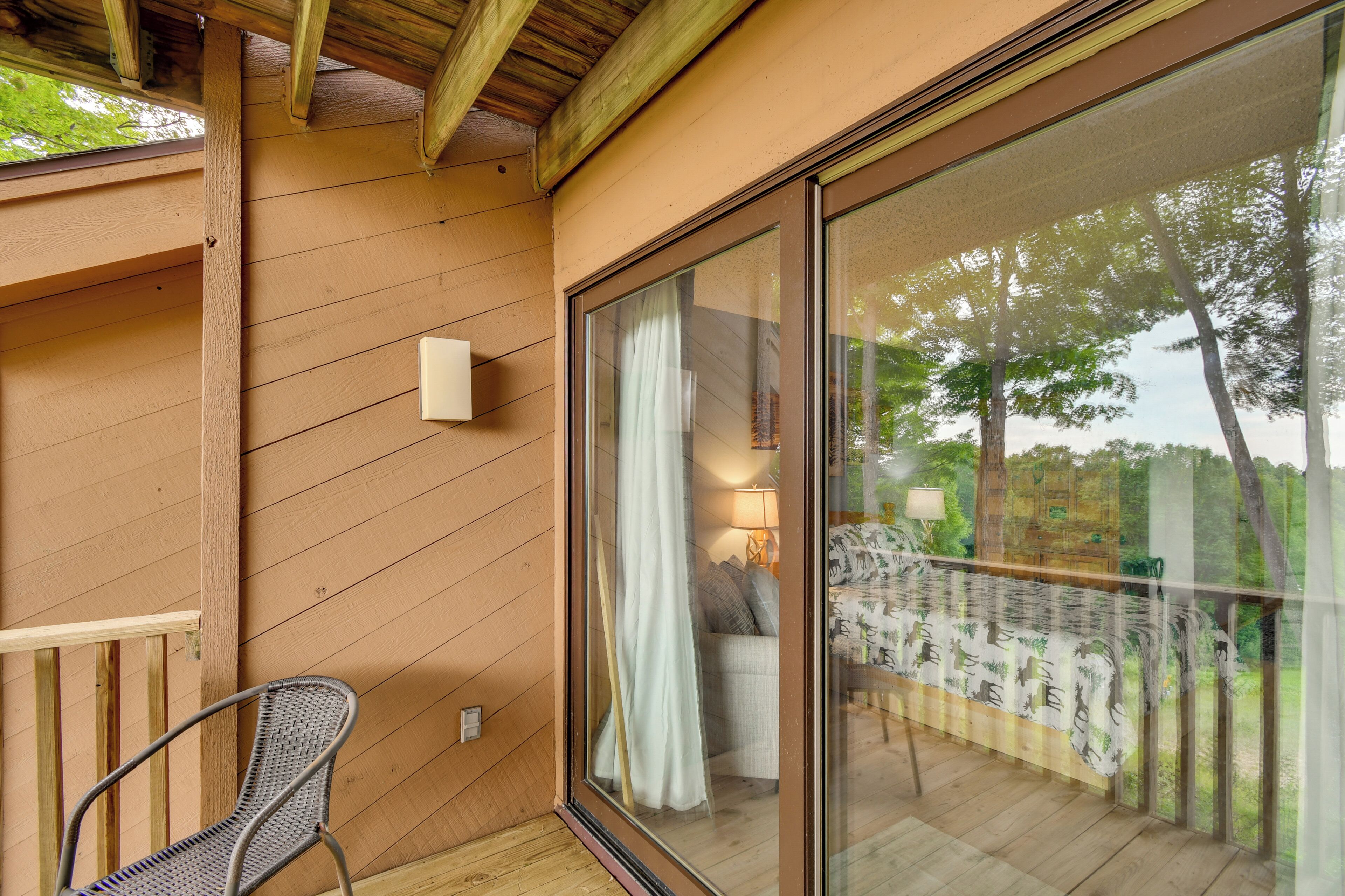 Une terrasse en bois avec une chaise, une porte vitrée donnant sur un balcon avec vue sur les arbres, et une lampe fixée au mur.
