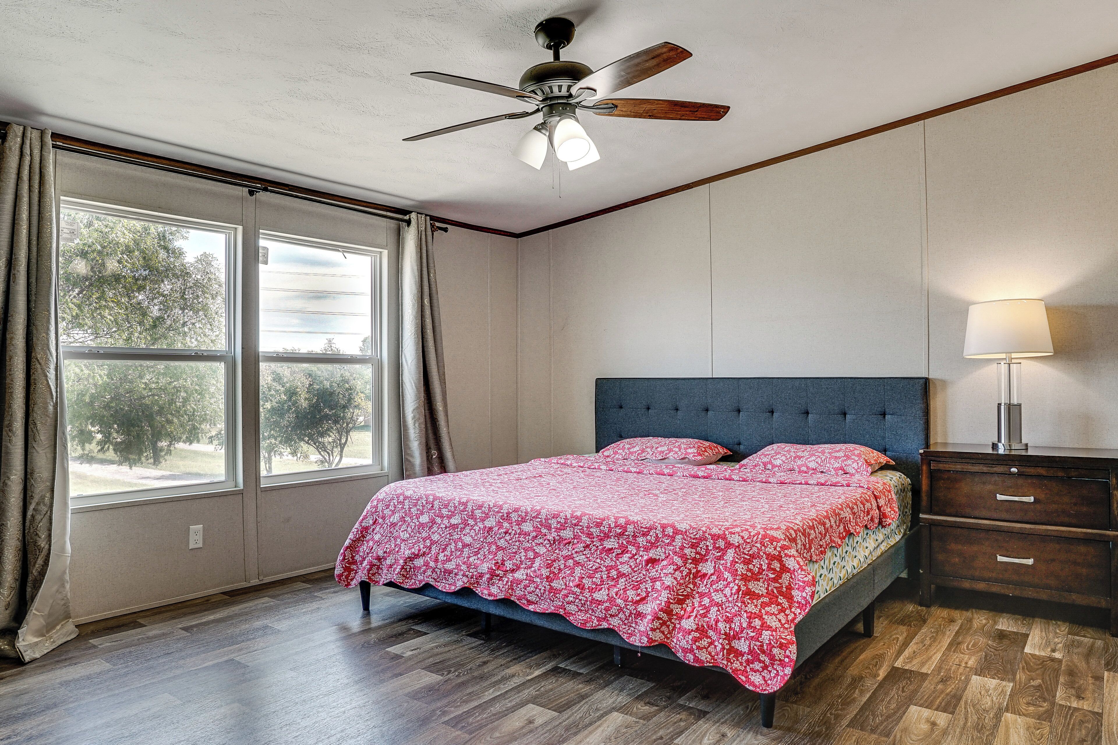Une chambre à coucher avec un lit, un ventilateur de plafond, une table de chevet, une lampe et une fenêtre avec des rideaux.