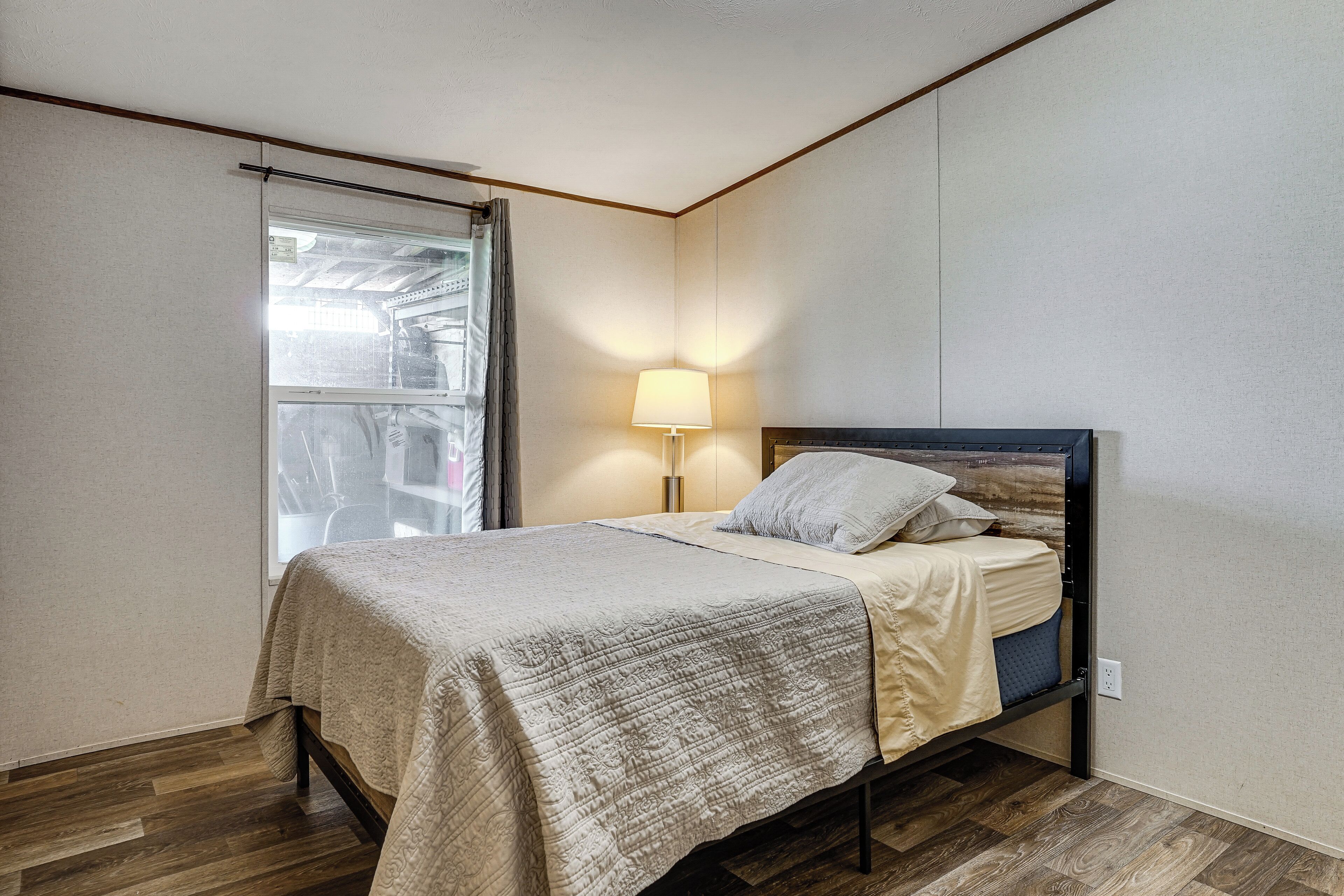 A bedroom with a bed, a window with curtains, a bedside lamp, and a wooden floor.