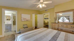 A bedroom with a bed, a ceiling fan, a dresser, a bench, and a door leading to a bathroom. - Jasmine Arbor - 2BR Apt in Coveted Historic Dtwn (Savannah)