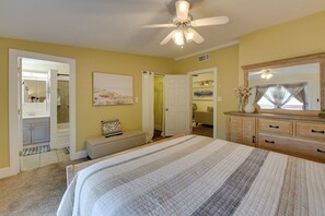 A bedroom with a bed, a ceiling fan, a dresser, a bench, and a door leading to a bathroom. - Jasmine Arbor - 2BR Apt in Coveted Historic Dtwn (Savannah)