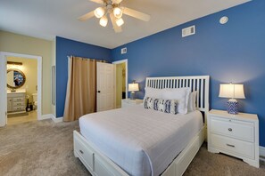 Une chambre à coucher avec un lit, des tables de chevet, un ventilateur de plafond et une salle de bain visible à travers une porte ouverte.